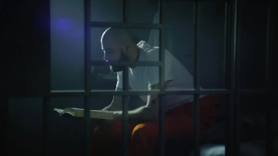 Male prisoner in orange uniform sits on bed in prison cell, reads Bible. Illegally convicted man serves imprisonment term in jail. View through metal bars. Detention center or correctional facility.