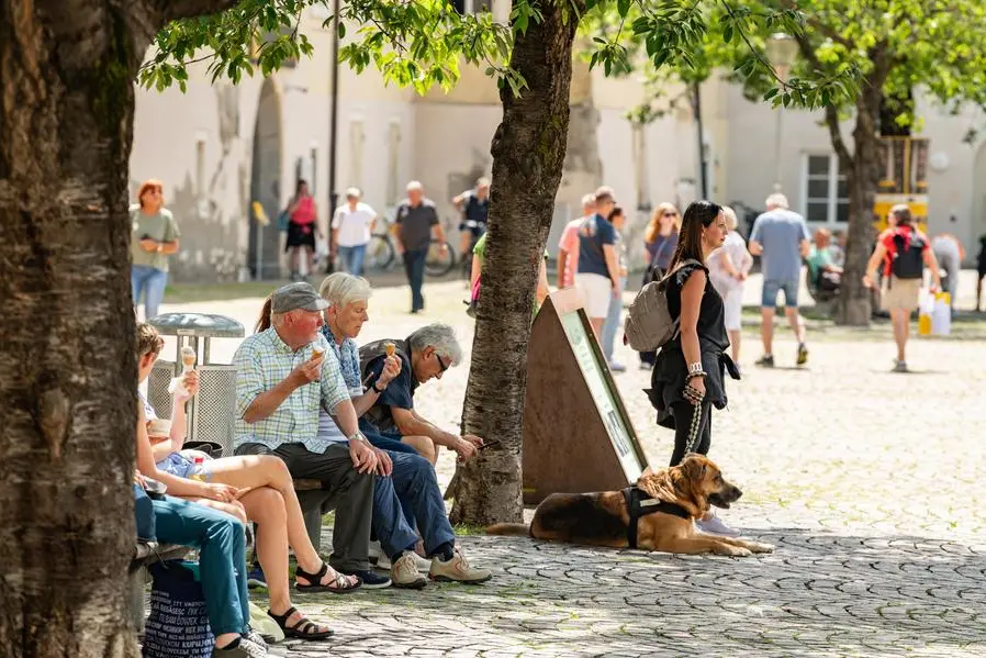 People of different age and gender eating an ice-cream on the grand-place, Piazza del Duomo Brixen (Bressanone) during summertime. Hot summer, people sitting in the shade. 20 July 2024, Brixen, Italy
