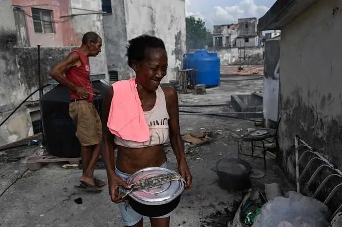 A woman smiles as she carries a pot of food during a blackout in Havana on May 13, 2026. Cuba blamed the United States for the \\\"particularly tense\\\" situation in its power grid on May 13, 2026, which has been plagued by prolonged blackouts, while Washington once again offered $100 million in aid to the island. (Photo by Yamil LAGE / AFP)