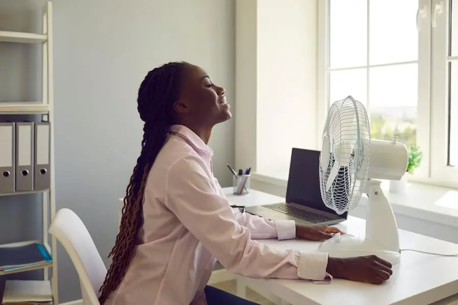 Young African American woman corporate worker, sitting at office table with laptop computer on hot summer day, switches on electric fan, feels refreshed and enjoys fresh cool air blowing in her face