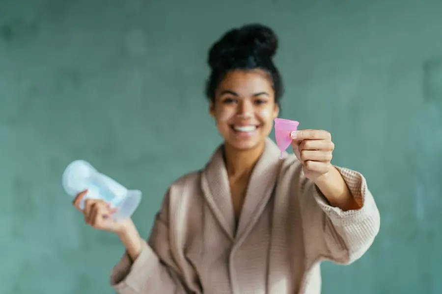 Cheerful energetic young woman recommending to use menstrual cup during critical days, holding hygiene pad in one hand on green bathroom background. eco friendly, zero waste, reuse concept.