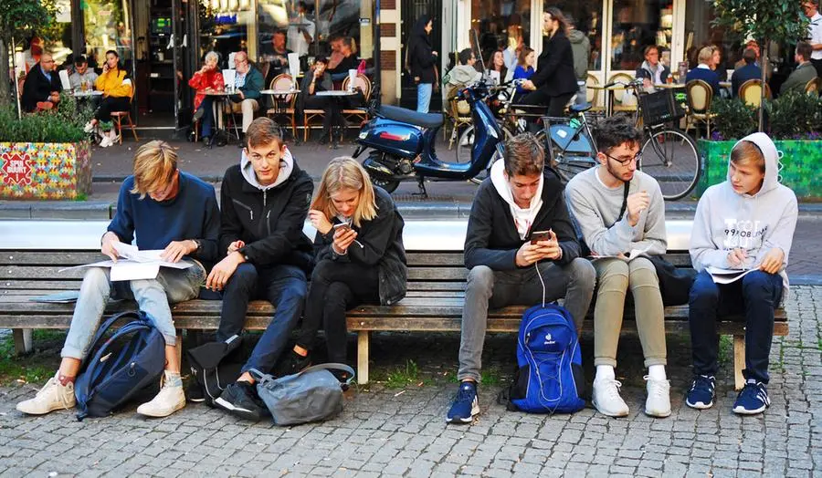 A group of secondary school boys sitting together on a publc square in the center of Amsterdam doing some school home work and playing togehter with their mobile phones.