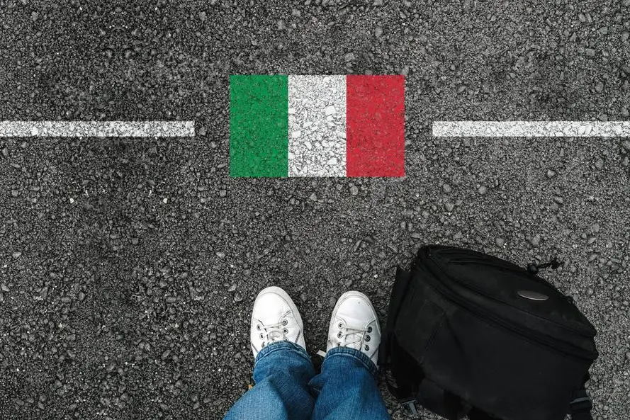 a man with a shoes and backpack is standing on asphalt next to flag of Italy and border
