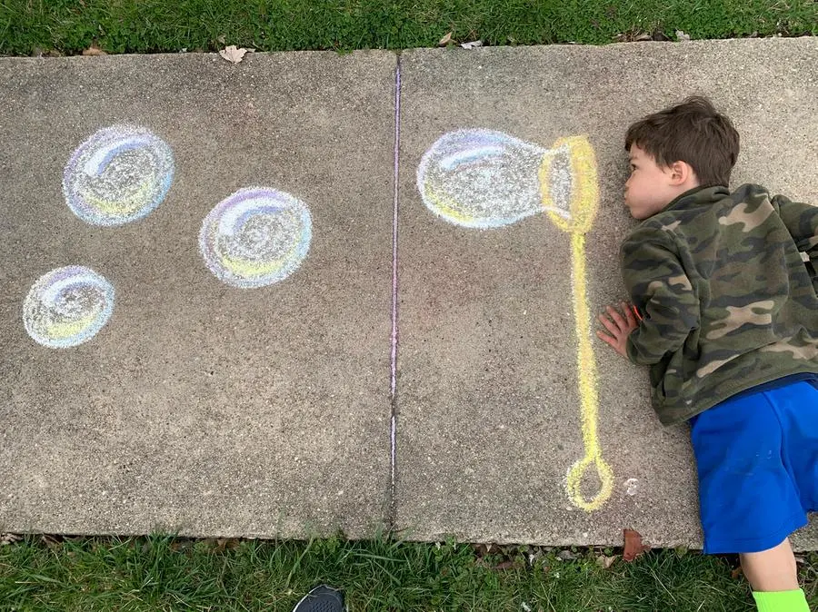 One young boy with green jacket and blue shorts lying on a sidewalk pretending to blow bubbles next to a chalk drawing of a wand and bubbles